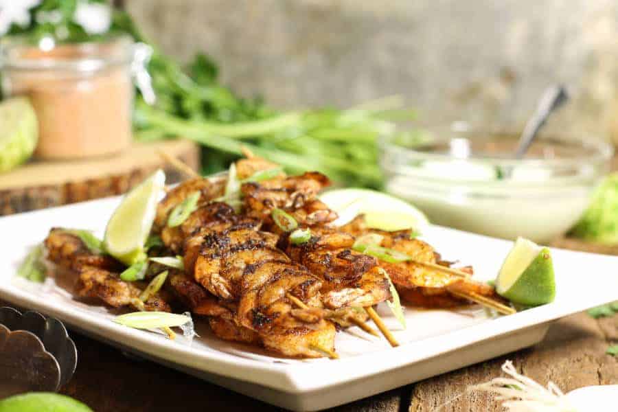 blackened shrimp skewers on a white platter surrounded by dip and various ingredients