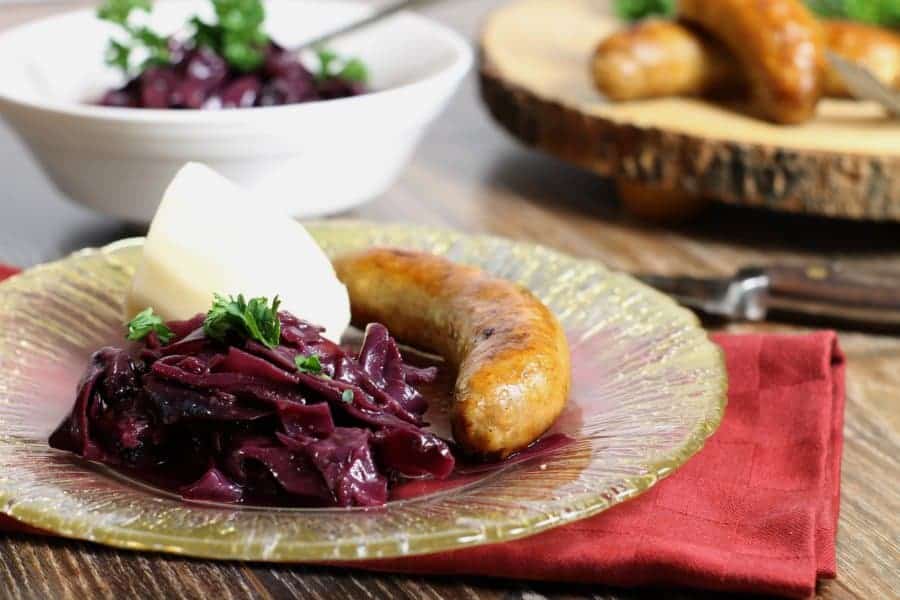 braised German red cabbage served on a fancy gold rimmed glass plate alongside sausages and boiled potato.