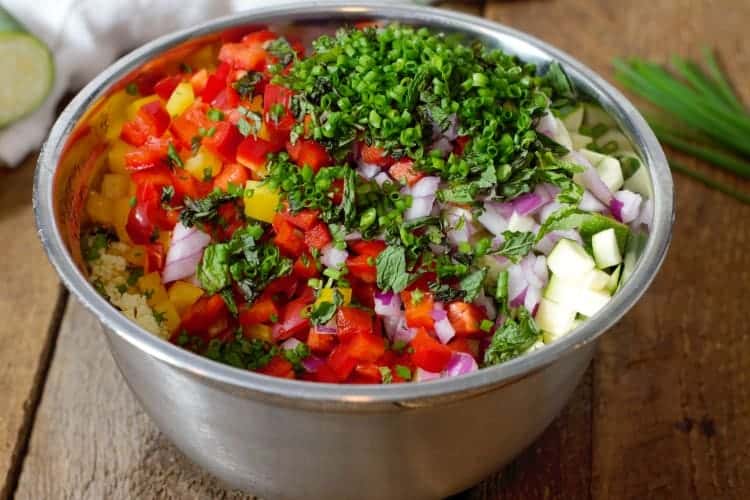 a stainless steel bowl of various vegetables and herbs which will be added to a Mediterranean Couscous Salad