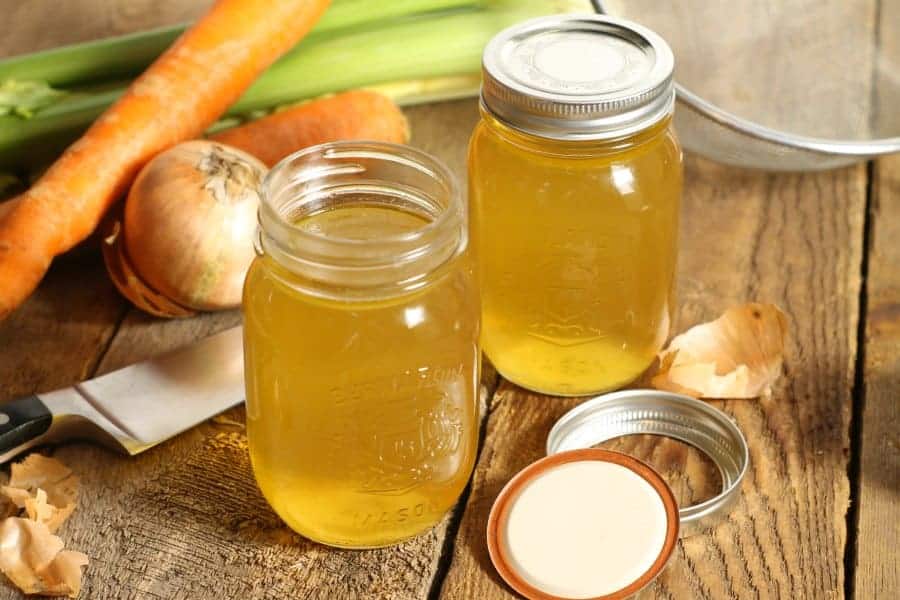 two mason jars on a wooden table filled with clear golden chicken stock. The jars are surrounded by various vegetables and a kitchen knife.