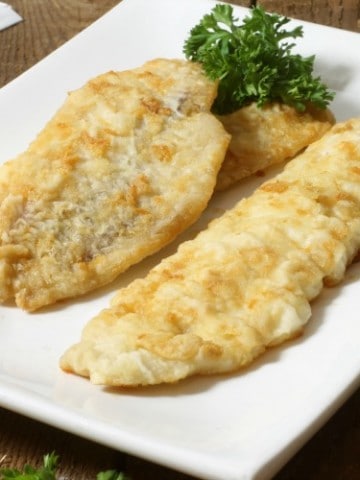 three pan fried pieces of haddock in a white plate with lemon wedges and parsley